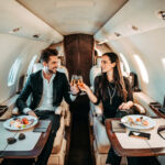 Rich couple making a toast with champagne glasses while eating canapes aboard a private jet