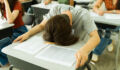 Overwhelmed high school student resting head on desk during difficult examination, surrounded by classmates