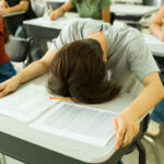 Overwhelmed high school student resting head on desk during difficult examination, surrounded by classmates
