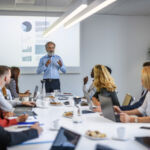 Bearded corporate professional explaining forecasting details with visual aid in presentation to executive team.