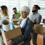 Large group of displeased business people carrying carton boxed with their belongings after being fired from their jobs. Focus is on senior woman.