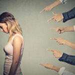 Concept of accusation guilty person girl. Side profile sad upset woman looking down many fingers pointing at her back isolated on grey office wall background. Human face expression emotion feeling