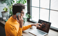 Young man at home office. He is sitting near window and using smart phone while working on laptop.