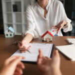 A woman is signing a document in front of a house model. The woman is wearing a white shirt and the house model is red