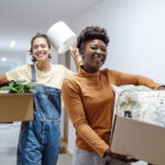 Two female friends carrying cardboard boxes with belongings and moving into campus
