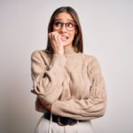 Young beautiful brunette businesswoman wearing casual sweater and glasses standing looking stressed and nervous with hands on mouth biting nails. Anxiety problem.