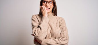 Young beautiful brunette businesswoman wearing casual sweater and glasses standing looking stressed and nervous with hands on mouth biting nails. Anxiety problem.