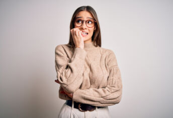 Young beautiful brunette businesswoman wearing casual sweater and glasses standing looking stressed and nervous with hands on mouth biting nails. Anxiety problem.