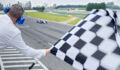 Rear view of auto race official waving chequered flag to blue Formula 1 car driving on track.