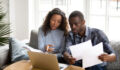 Serious African American couple discussing paper documents, sitting together on couch at home, man and woman checking bills, bank account balance, terms of contract, mortgage, loan agreement