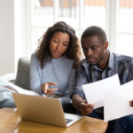 Serious African American couple discussing paper documents, sitting together on couch at home, man and woman checking bills, bank account balance, terms of contract, mortgage, loan agreement