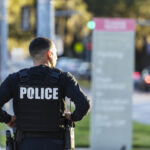 Rear view of an Hispanic police officer patrolling an urban area on foot. He is walking on a sidewalk away from the camera.