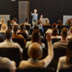 Happy mature speaker giving a speech in front of people at convention center while some of them wants to ask a question.