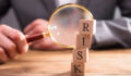 Close-up Of Wooden Blocks With Risk Word In Front Of Businessperson's Hand Holding Magnifying Glass
