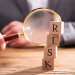 Close-up Of Wooden Blocks With Risk Word In Front Of Businessperson's Hand Holding Magnifying Glass