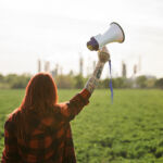 Rear view of young woman activist with megaphone standing outdoors by oil refinery, protesting.
