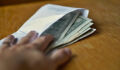 Male hand opening a white envelope full of American Dollars (USD, US Dollars) on the wooden table as a symbol of cash transfer, money laundering or bribery