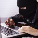 Masked theif wearing all black steals computer secrets or commits identity theft on an office or home laptop computer. Cyper crimes concept.