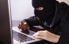 Masked theif wearing all black steals computer secrets or commits identity theft on an office or home laptop computer. Cyper crimes concept.