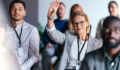 Focused businesswoman raises hand during a professional event, indicating engagement and participation. Diverse group of people in a coworking setting, highlighting teamwork and collaboration.