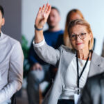 SARs and RIAs following new FAQs Focused businesswoman raises hand during a professional event, indicating engagement and participation. Diverse group of people in a coworking setting, highlighting teamwork and collaboration.