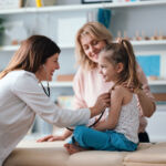 A friendly pediatrician performs a health examination for a happy young girl, observed by her caring mother. The image conveys family care, child health, and professional medical service.