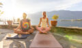Two people practicing yoga meditation on mats beside a tranquil lake. Peaceful morning wellness routine under the rising sun. Outdoor mindfulness activity amidst nature.