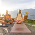Two people practicing yoga meditation on mats beside a tranquil lake. Peaceful morning wellness routine under the rising sun. Outdoor mindfulness activity amidst nature.