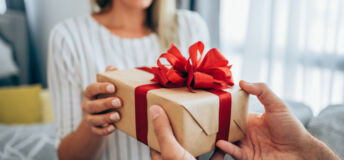 Cropped shot of a loving husband giving his wife a gift. Boyfriend surprise his beautiful girlfriend with present while she is sitting on the sofa in the living room at home. Focus is on the gift.