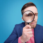 Serious young male detective holding magnifying glass by right eye while standing in front of camera in isolation