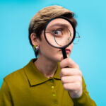 A confident young woman holds a magnifying glass, focusing on its details, dressed stylishly in a casual outfit, set against a vibrant blue background.
