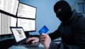 Man wearing balaclava and holding credit card while using laptop at desk