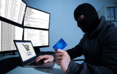 Man wearing balaclava and holding credit card while using laptop at desk