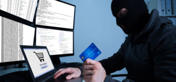 Man wearing balaclava and holding credit card while using laptop at desk