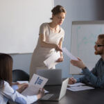 Broker-Dealers: SEC focusing on customer protection, P&Ps Millennial businesswoman give handout materials to work team members during flipchart presentation, female speaker or coach share documents to workers, presenter hand papers to consider or analyze