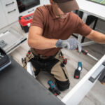 Caucasian Male Worker Installing the Handle to the Kitchen Cabinet Door Using Screwdriver. Modern New Kitchen and Other Professional Tools for Installing and Repairing Furniture in the Background.