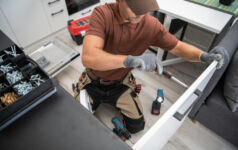 Caucasian Male Worker Installing the Handle to the Kitchen Cabinet Door Using Screwdriver. Modern New Kitchen and Other Professional Tools for Installing and Repairing Furniture in the Background.