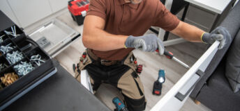Caucasian Male Worker Installing the Handle to the Kitchen Cabinet Door Using Screwdriver. Modern New Kitchen and Other Professional Tools for Installing and Repairing Furniture in the Background.