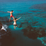 Young active man and woman diving from high cliff into tropical island blue sea water