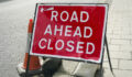 A sign reading “Road Ahead Closed” weighted down by sand bags, half on the pavement and half on the road in an English town.