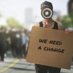 public protest and riots in the city streets. woman with megaphone and cardboard poster in hands on protesters crowd background. democracy concept