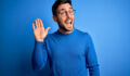 Young handsome man with beard wearing casual sweater and glasses over blue background Waiving saying hello happy and smiling, friendly welcome gesture