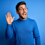 Young handsome man with beard wearing casual sweater and glasses over blue background Waiving saying hello happy and smiling, friendly welcome gesture