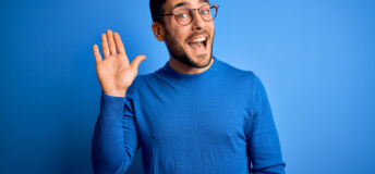 Young handsome man with beard wearing casual sweater and glasses over blue background Waiving saying hello happy and smiling, friendly welcome gesture
