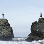 A businesswoman tries to communicate through a megaphone with a businessman as they stand on separate rocky bluffs high above the ocean waves.