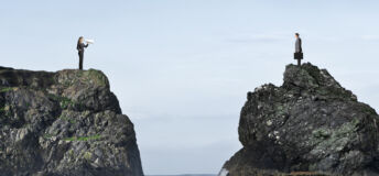 A businesswoman tries to communicate through a megaphone with a businessman as they stand on separate rocky bluffs high above the ocean waves.
