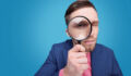 Serious young male detective holding magnifying glass by right eye while standing in front of camera in isolation