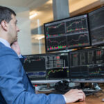 Male stock broker trading online watching charts and data analyses on multiple computer screens.