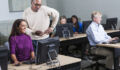 Continuing education: multi-ethnic adult students in a classroom working on desktop computers. The focus is on the two people in the foreground, a mid adult woman wearing a purple shirt sitting at a computer, and a young African American man wearing glasses, standing beside her, looking at her screen. Three other people are working in the background.