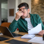 Anxious male financial advisor rubbing eyes while working over laptop and analyzing reports at desk